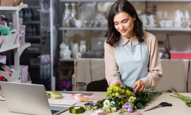kwiaciarka w błękitnym fartuchu układająca bukiet kwiatów na blacie, na którym leży również otwarty laptop