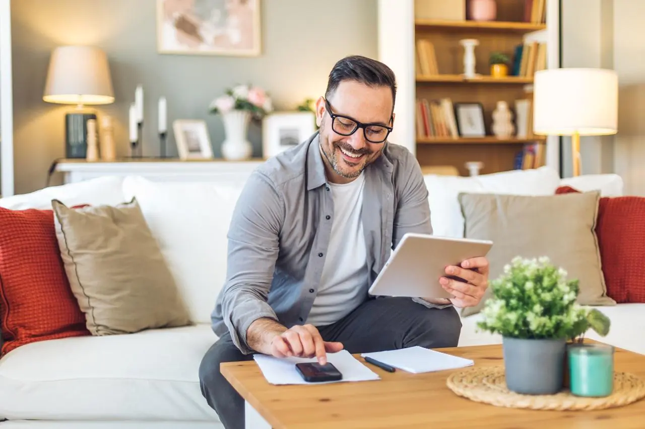 Uśmiechający się mężczyzna, siedzący w salonie i obliczający na kalkulatorze podatek Belki w 2026 roku.