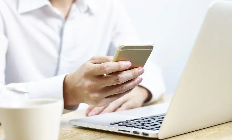 kobieta w białej koszuli siedząca przed laptopem z telefonem w dłoni