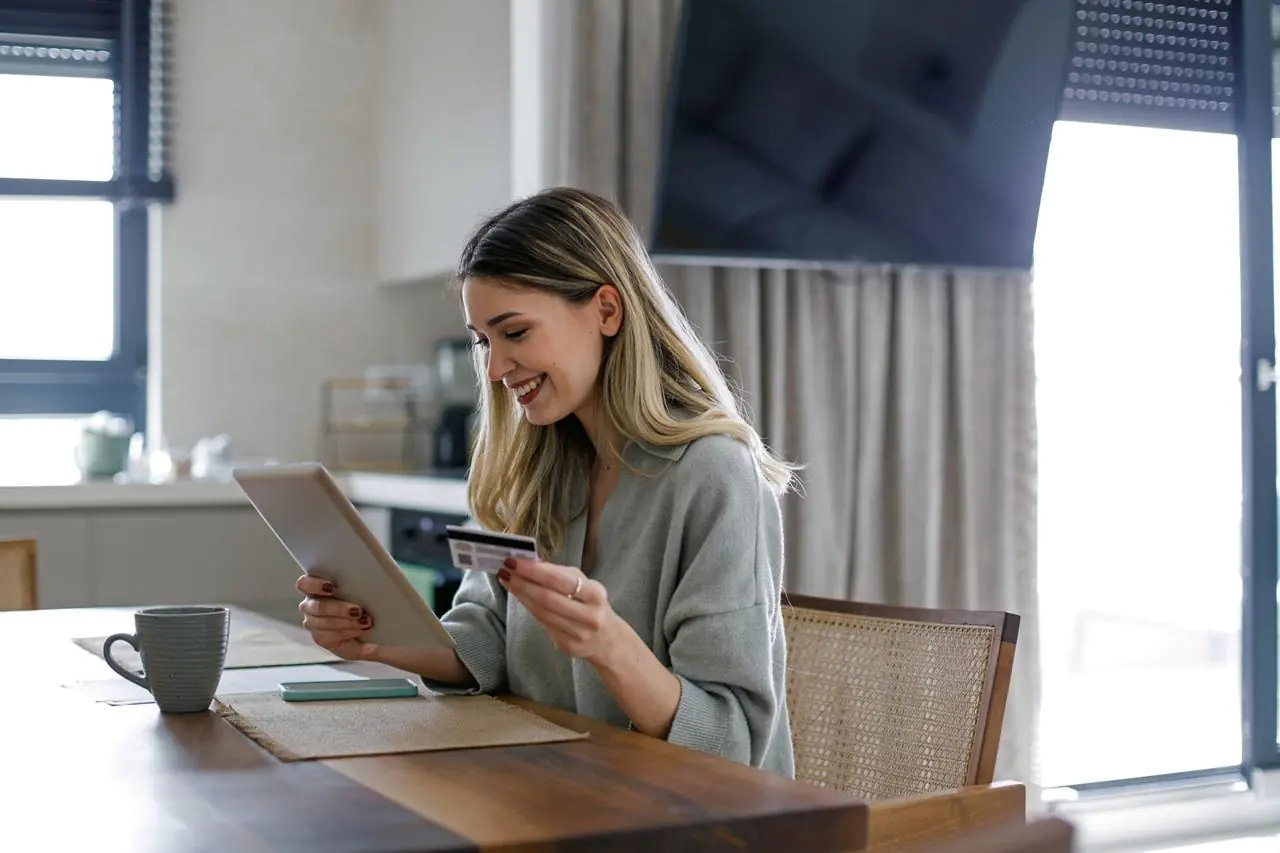 Młoda kobieta siedząca przy stole z tabletem i kartą, która sprawdza w internecie, co się bardziej opłaca - karta kredytowa czy kredyt?