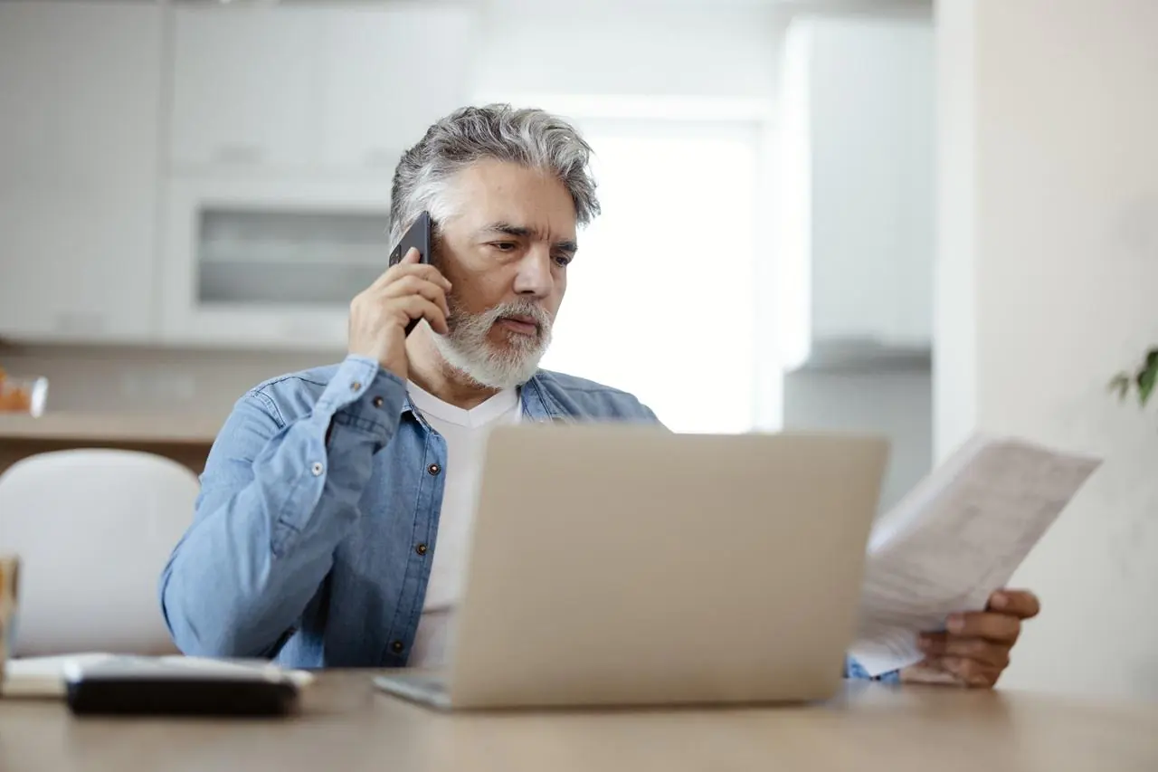 Starszy mężczyzna siedzący przed laptopem i dzwoniący na infolinię banku w celu zastrzeżenia karty płatniczej