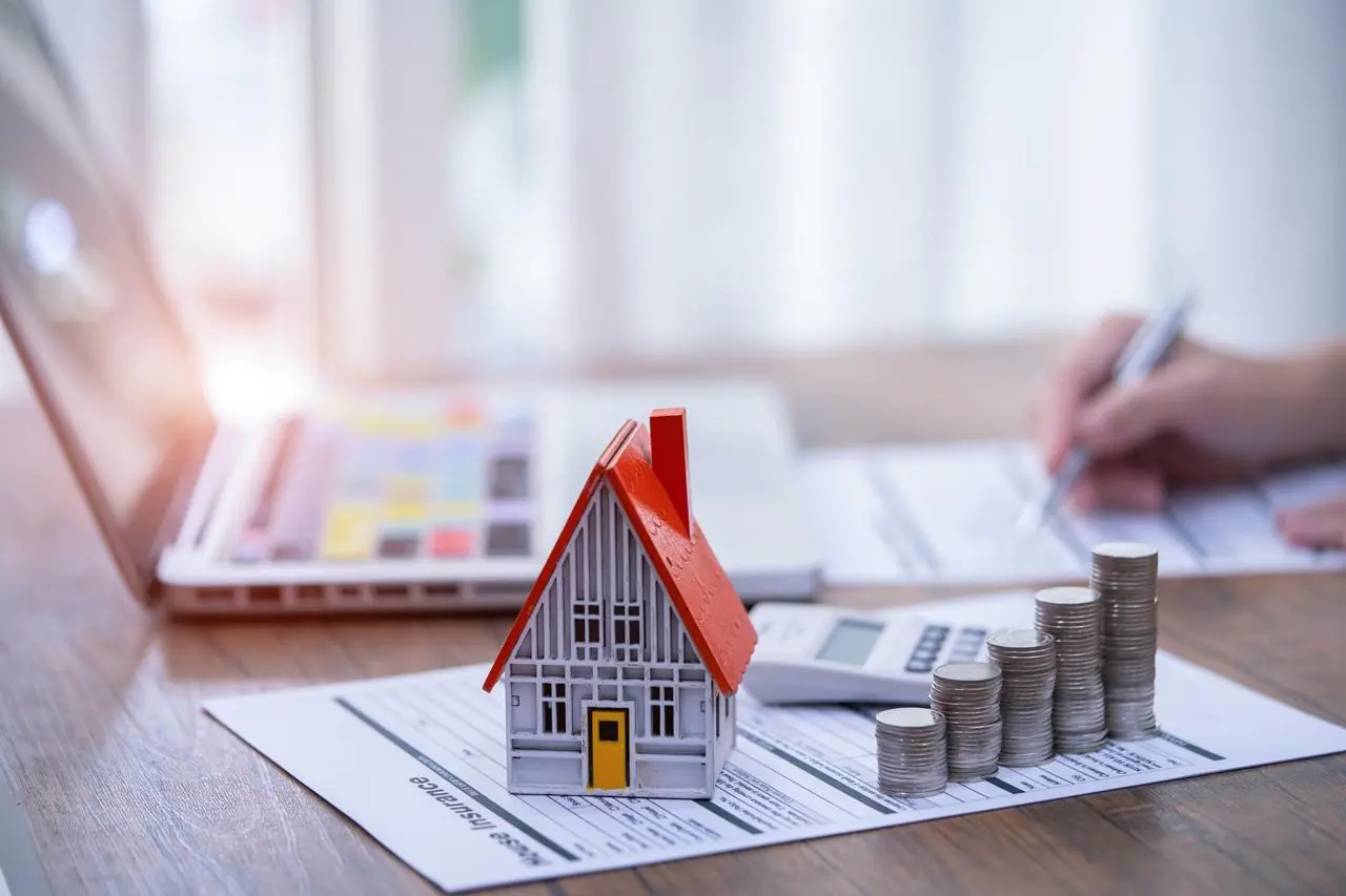 Makieta domu jednorodzinnego, obok rosnące słupki z monet, w tle kobieta obliczająca koszt ubezpieczenia pomostowego.