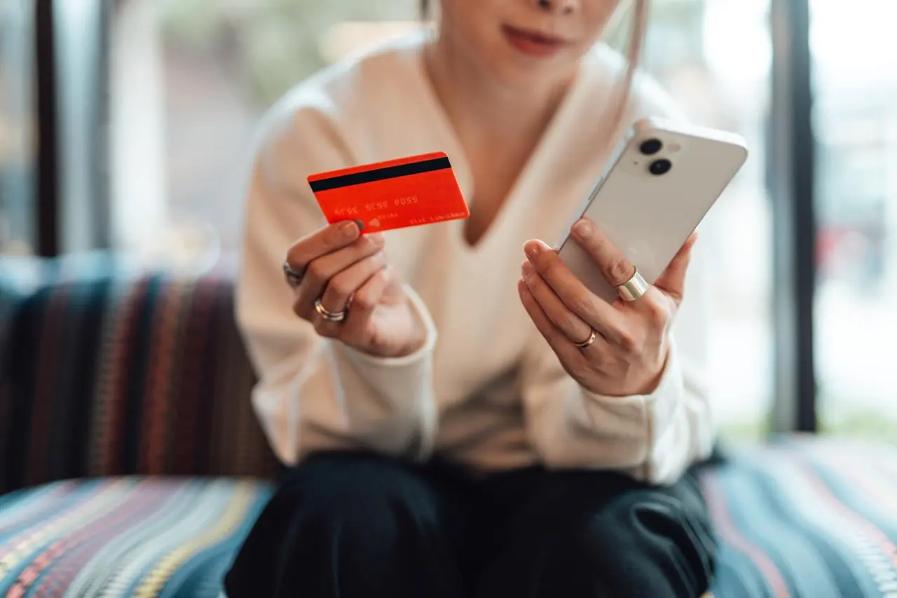 Kobieta w białej bluzce trzyma w jednej dłoni kartę kredytową, a w drugiej telefon
