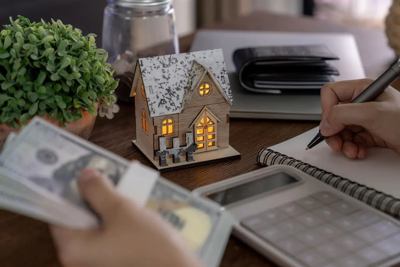 Makieta domu, kalkulator, portfel i otwarty notes, w którym kobieta chce obliczyć, ile trzeba zarabiać, żeby dostać kredyt hipoteczny.