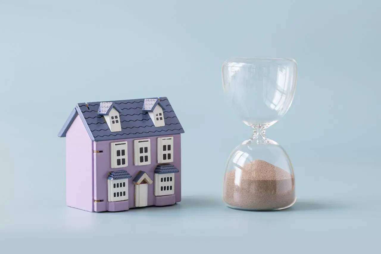 Fioletowy model domu jednorodzinnego stojący obok klepsydry na błękitnym tle.