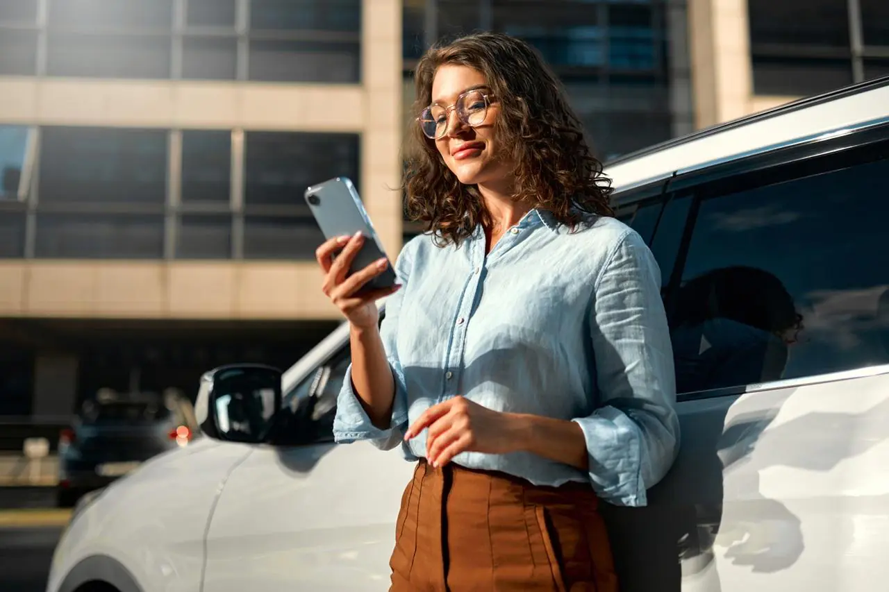 zadowolona kobieta opierająca się o samochód i sprawdzająca w smartfonie, jak wygląda jej historia kredytowa