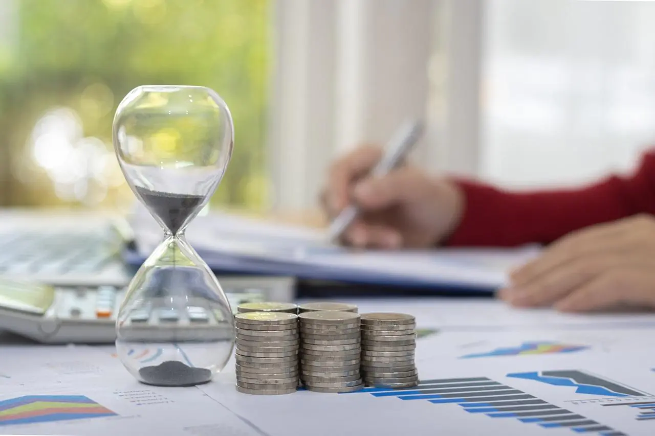 Szklana klepsydra i stosy monet, a w tle kobieta sprawdzająca w dokumentach, czy jest możliwy kredyt gotówkowy na 15 lat.
