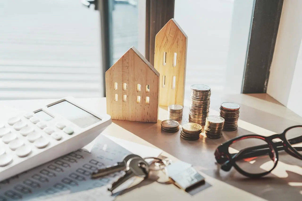 Drewniane figurki domów stojące na biurku obok kalkulatora, kluczy, słupków monet i okularów.