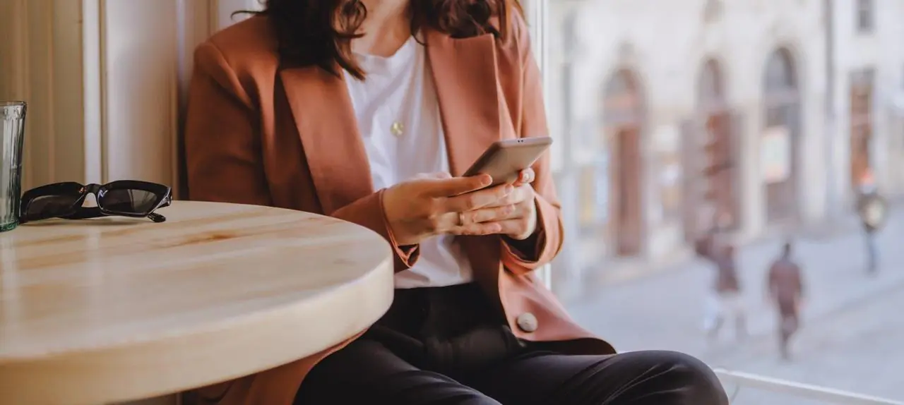 Kobieta w brązowej marynarce trzyma w rękach telefon i robi przelew BLIKIEM