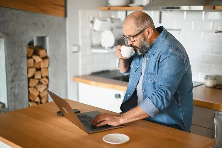 mężczyzna stojący z kawą w kuchni i sprawdzający na laptopie ranking pożyczek 1100 zł