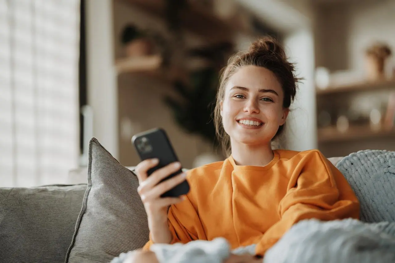 Młoda kobieta w pomarańczowej bluzce siedzi na kanapie i trzyma w dłoni telefon