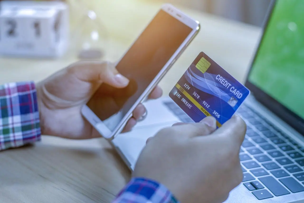 Mężczyzna trzyma w dłoniach kartę kredytową i telefon siedząc przed laptopem