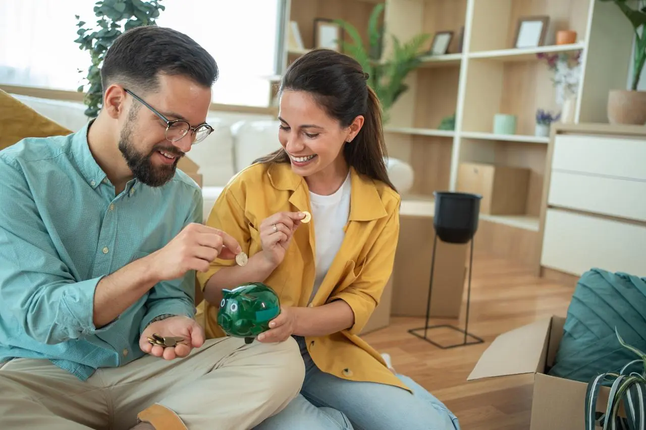 Młoda, zadowolona para siedzi w salonie i wrzuca monety do zielonej świnki-skarbonki.