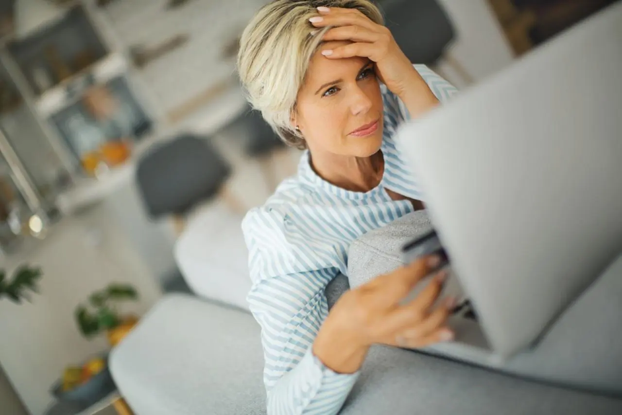 Wyraźnie zmartwiona kobieta trzyma w dłoniach kartę bankomatową i patrzy w ekran laptopa