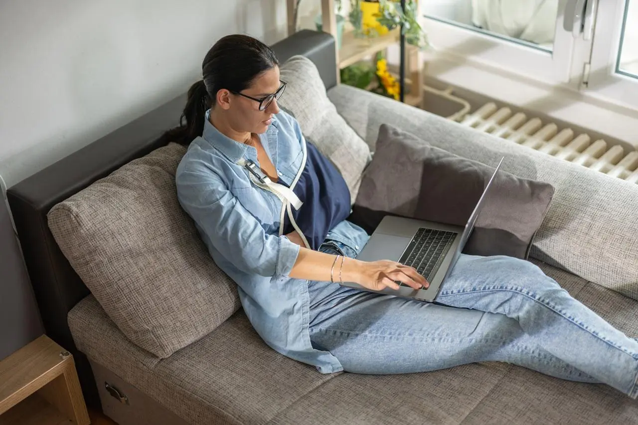 Kobieta ze złamaną ręką siedząca na sofie i sprawdzająca na laptopie warunki ubezpieczenia kredytu gotówkowego