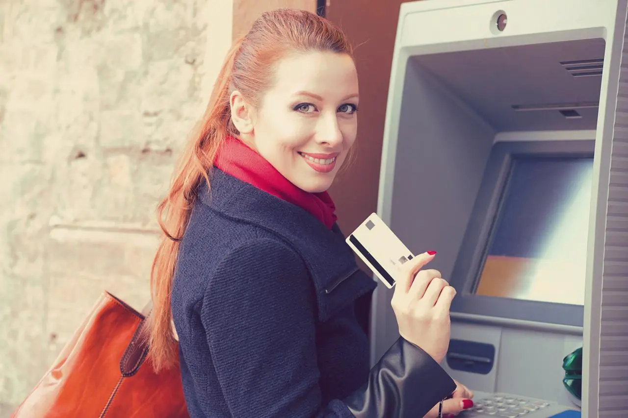 Kobieta z rudymi włosami stoi przed bankomatem z kartą płatniczą w ręku