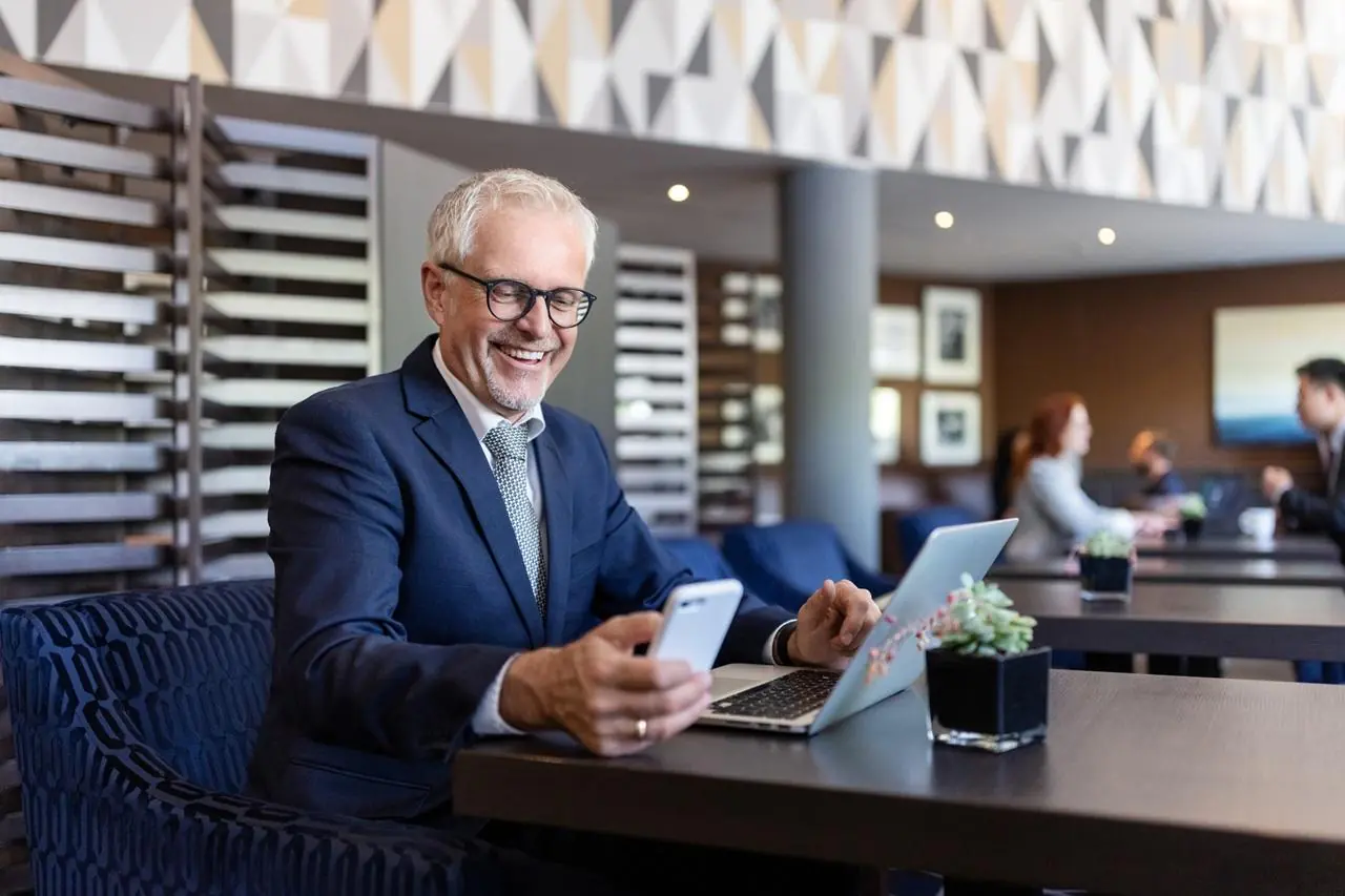 Starszy, zadowolony biznesmen z laptopem i telefonem komórkowym sprawdza w internecie najlepsze lokaty bankowe na 6 miesięcy.