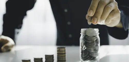 mężczyzna w granatowej koszuli wrzucający monetę do słoika pełnego oszczędności