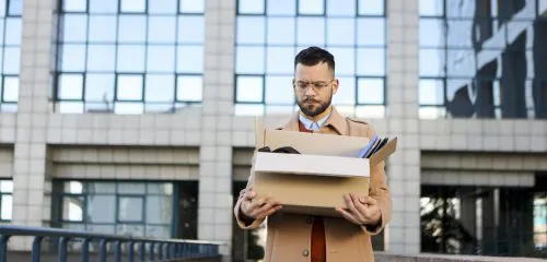 Smutny mężczyzna wyrzucony z pracy stoi z kartonem w rękach przed budynkiem firmy.