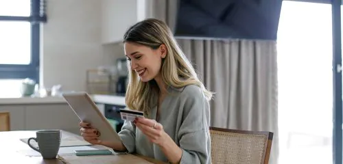 Młoda kobieta siedząca przy stole z tabletem i kartą, która sprawdza w internecie, co się bardziej opłaca - karta kredytowa czy kredyt?