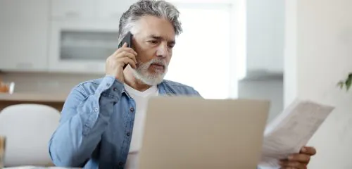 Starszy mężczyzna siedzący przed laptopem i dzwoniący na infolinię banku w celu zastrzeżenia karty płatniczej