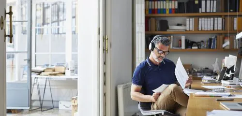 Mężczyzna z słuchawkami na uszach, siedzący w mieszkaniu i sprawdzający w dokumentach numer rachunku bankowego