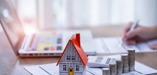 Makieta domu jednorodzinnego, obok rosnące słupki z monet, w tle kobieta obliczająca koszt ubezpieczenia pomostowego.