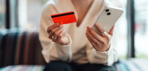 Kobieta w białej bluzce trzyma w jednej dłoni kartę kredytową, a w drugiej telefon