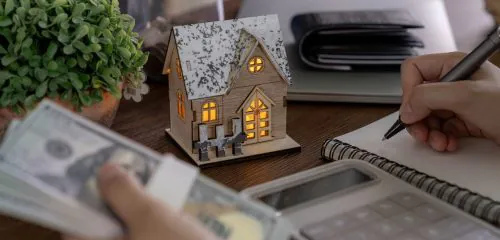 Makieta domu, kalkulator, portfel i otwarty notes, w którym kobieta chce obliczyć, ile trzeba zarabiać, żeby dostać kredyt hipoteczny.
