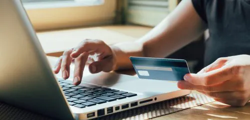 Zbliżenie na męskie dłonie. W jednej z nich karta, a druga na klawiaturze laptopa. Mężczyzna spłaca kartę kredytową w okresie bezodsetkowym,