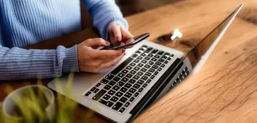 Młoda kobieta w niebieskim swetrze siedzi przed laptopem ze smarfonem w rękach