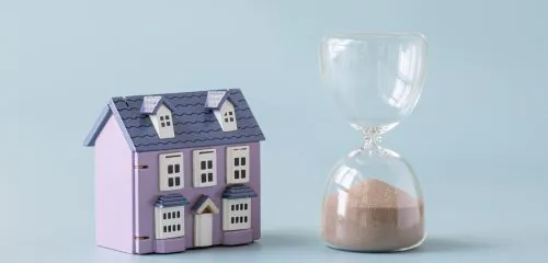 Fioletowy model domu jednorodzinnego stojący obok klepsydry na błękitnym tle.