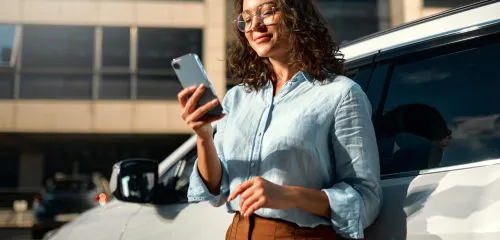 zadowolona kobieta opierająca się o samochód i sprawdzająca w smartfonie, jak wygląda jej historia kredytowa