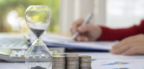 Szklana klepsydra i stosy monet, a w tle kobieta sprawdzająca w dokumentach, czy jest możliwy kredyt gotówkowy na 15 lat.