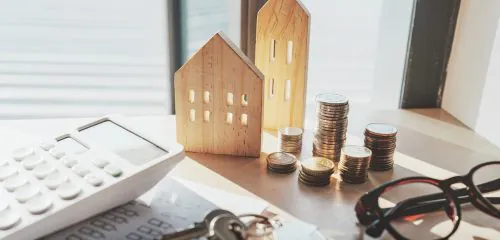 Drewniane figurki domów stojące na biurku obok kalkulatora, kluczy, słupków monet i okularów.