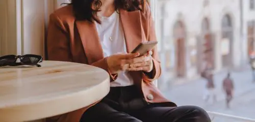 Kobieta w brązowej marynarce trzyma w rękach telefon i robi przelew BLIKIEM