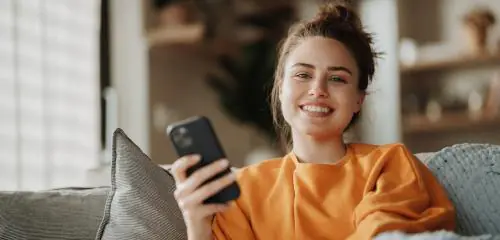 Młoda kobieta w pomarańczowej bluzce siedzi na kanapie i trzyma w dłoni telefon