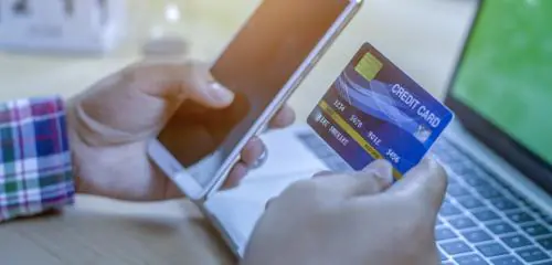 Mężczyzna trzyma w dłoniach kartę kredytową i telefon siedząc przed laptopem