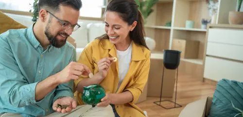 Młoda, zadowolona para siedzi w salonie i wrzuca monety do zielonej świnki-skarbonki.