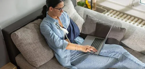 Kobieta ze złamaną ręką siedząca na sofie i sprawdzająca na laptopie warunki ubezpieczenia kredytu gotówkowego