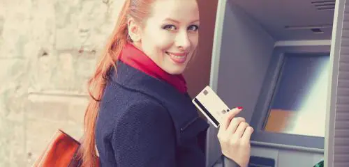Kobieta z rudymi włosami stoi przed bankomatem z kartą płatniczą w ręku