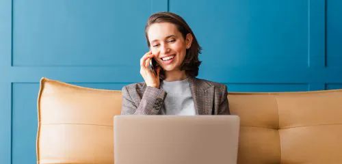 Kobieta siedząca na sofie, rozmawiająca przez telefon i sprawdzająca na laptopie ranking najlepszych lokat bankowych na 6 miesięcy