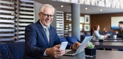 Starszy, zadowolony biznesmen z laptopem i telefonem komórkowym sprawdza w internecie najlepsze lokaty bankowe na 6 miesięcy.