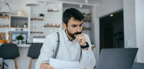 mężczyzna w białej koszuli siedzący przy biurku z umową i sprawdzający na laptopie kredyt na spłatę chwilówek