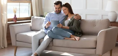 Młode małżeństwo siedzące na sofie i składające przez tablet wniosek online o dopisanie współwłaściciela do konta bankowego