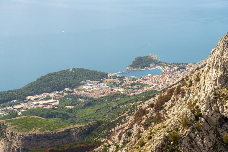 Panorama miasta Makarska u podnóża gór Biokovo. Na pierwszym planie widoczne są skaliste zbocza, poniżej zabudowa miejska, port i morze z niewielkimi statkami.