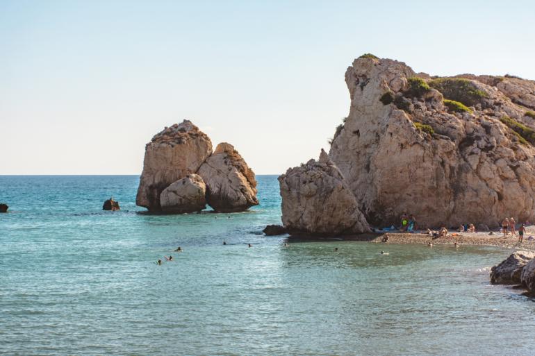 Skała Afrodyty w Pafos na Cyprze. Na zdjęciu widać duże wapienne formacje skalne wyrastające z błękitnego morza, przy których kąpią się ludzie. Wzdłuż brzegu leży wąska, kamienista plaża z turystami odpoczywającymi w słońcu.