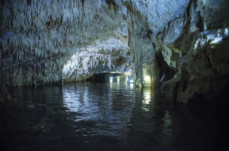Wnętrze Jaskiń Diros na południu Peloponezu. Widać podziemne korytarze częściowo zalane wodą, ozdobione naturalnymi formacjami skalnymi. Delikatne światło lamp podkreśla strukturę skał i nadaje miejscu tajemniczy klimat.