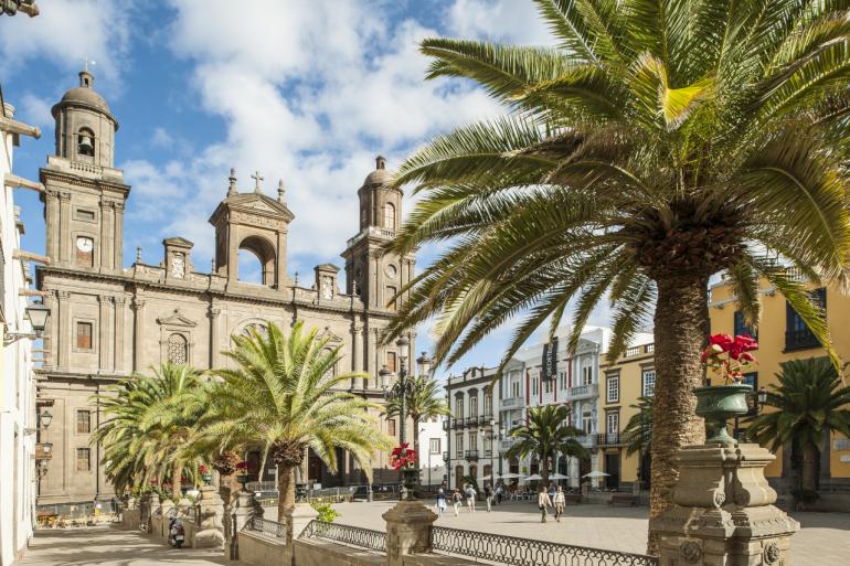 Katedra Santa Ana w Las Palmas de Gran Canaria otoczona palmami, z widocznym placem i spacerującymi turystami w słoneczny dzień.