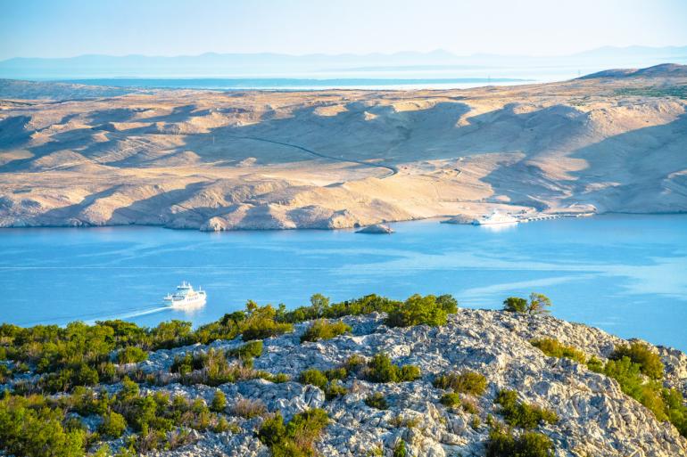 Zdjęcie przedstawia charakterystyczny, suchy i kamienisty krajobraz wyspy Pag w Chorwacji. W centrum widoczna jest zatoka z turkusowo-niebieską wodą, po której płynie biały prom. W tle rozciągają się złociste wzgórza i górzysty horyzont, a na pierwszym planie rosną niskie krzewy i skały, typowe dla tej części Dalmacji.